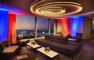 A modern lounge with a sofa, armchairs, and a coffee table. Large windows offer a city view.