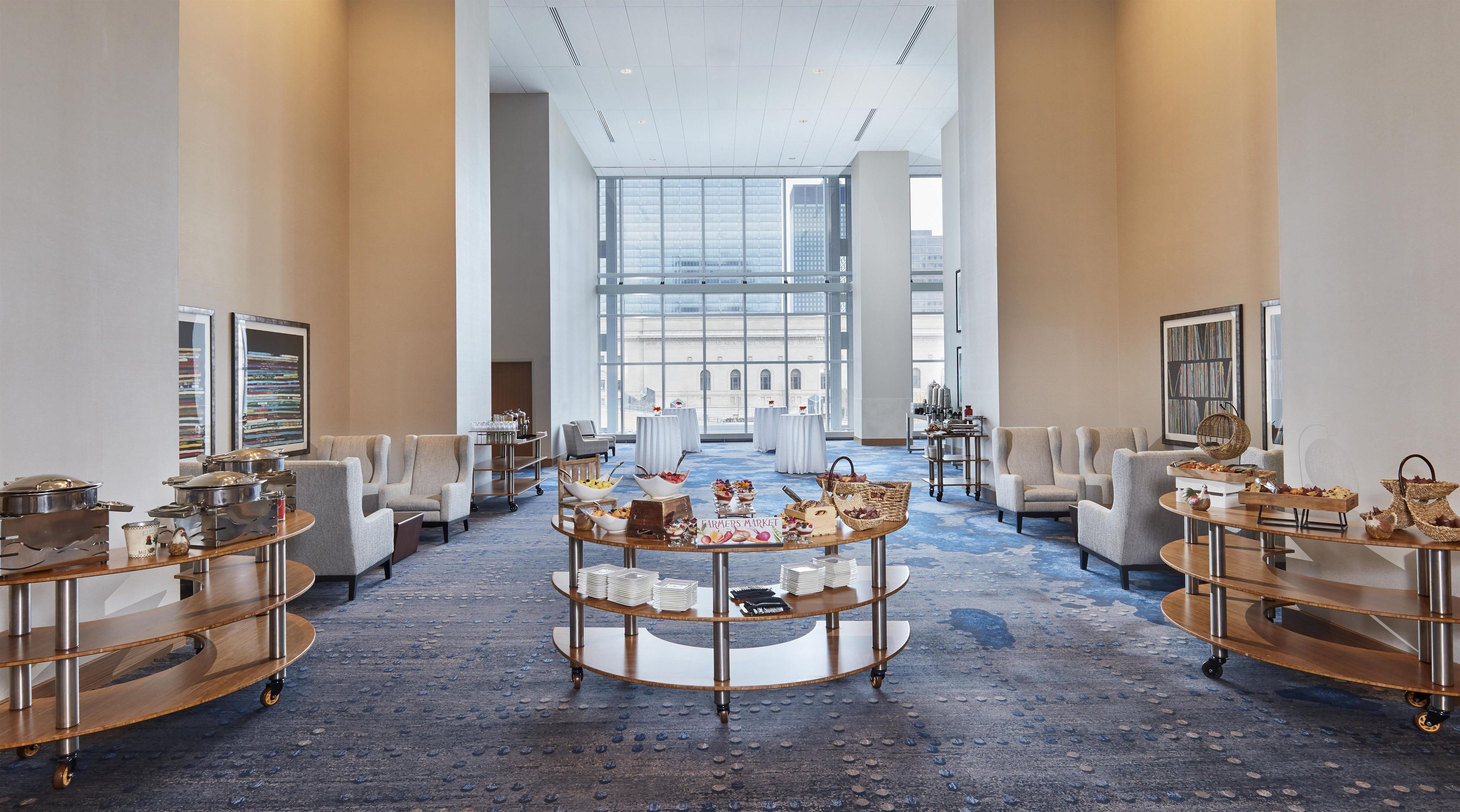 A spacious hotel lobby with a central round table displaying a farmers market sign, surrounded by seating and buffet carts.
