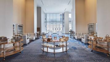 A spacious hotel lobby with a central round table displaying a farmers market sign, surrounded by seating and buffet carts.