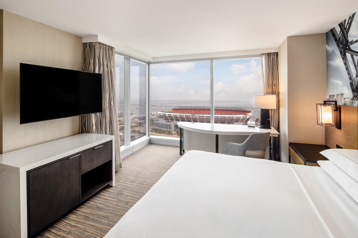 Ein Hotelzimmer mit einem großen Bett, einem flachbildfernseher an der Wand, einem Schreibtisch mit Stuhl und einem großen Fenster mit Blick auf ein Stadion.