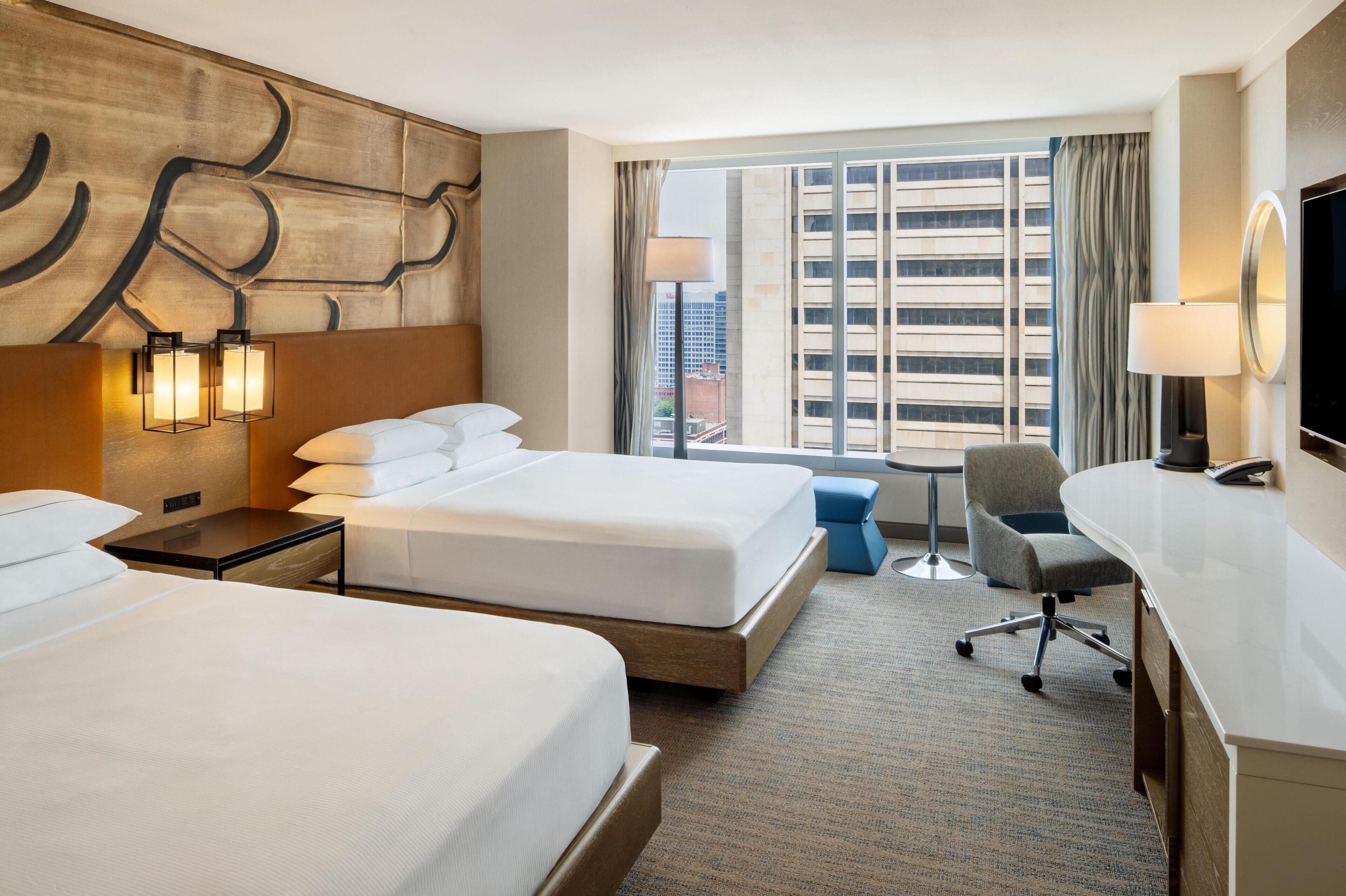Een hotelkamer met twee bedden, een bureau, een stoel en een groot raam met uitzicht op de stad.