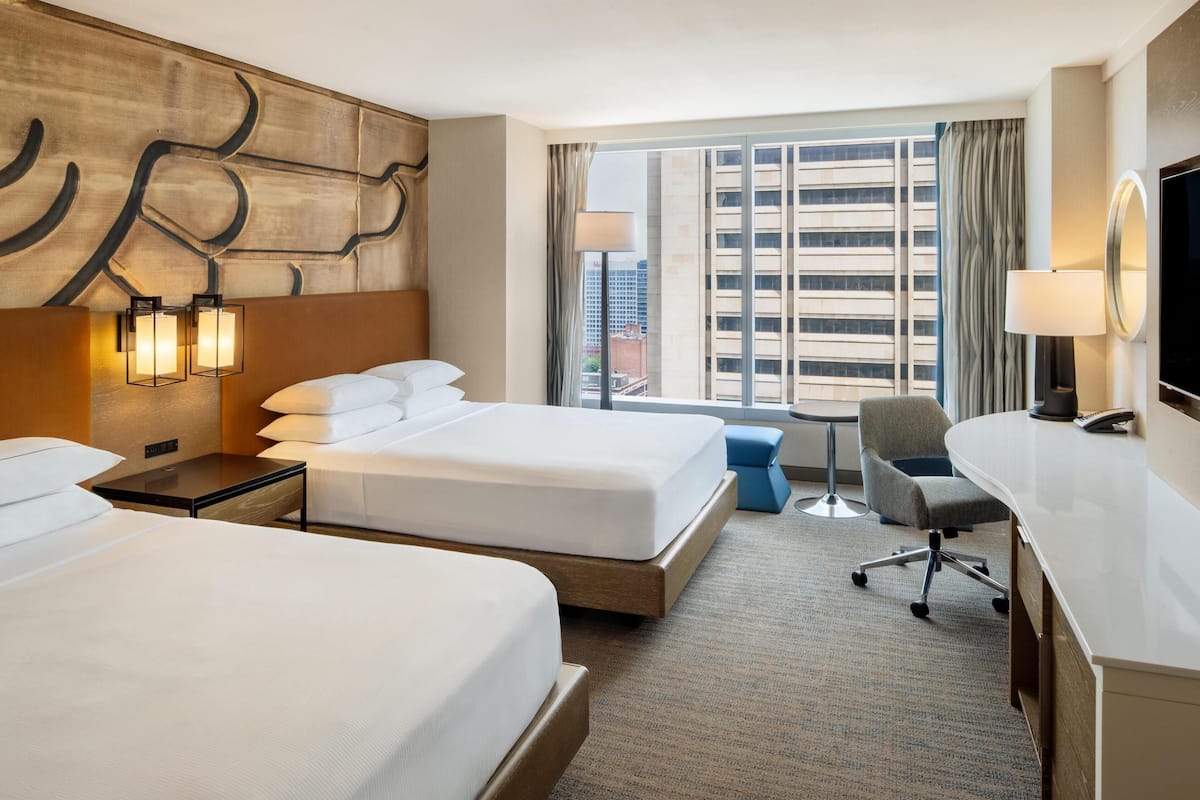 A hotel room with two beds, a desk, a chair, and a large window with a city view.
