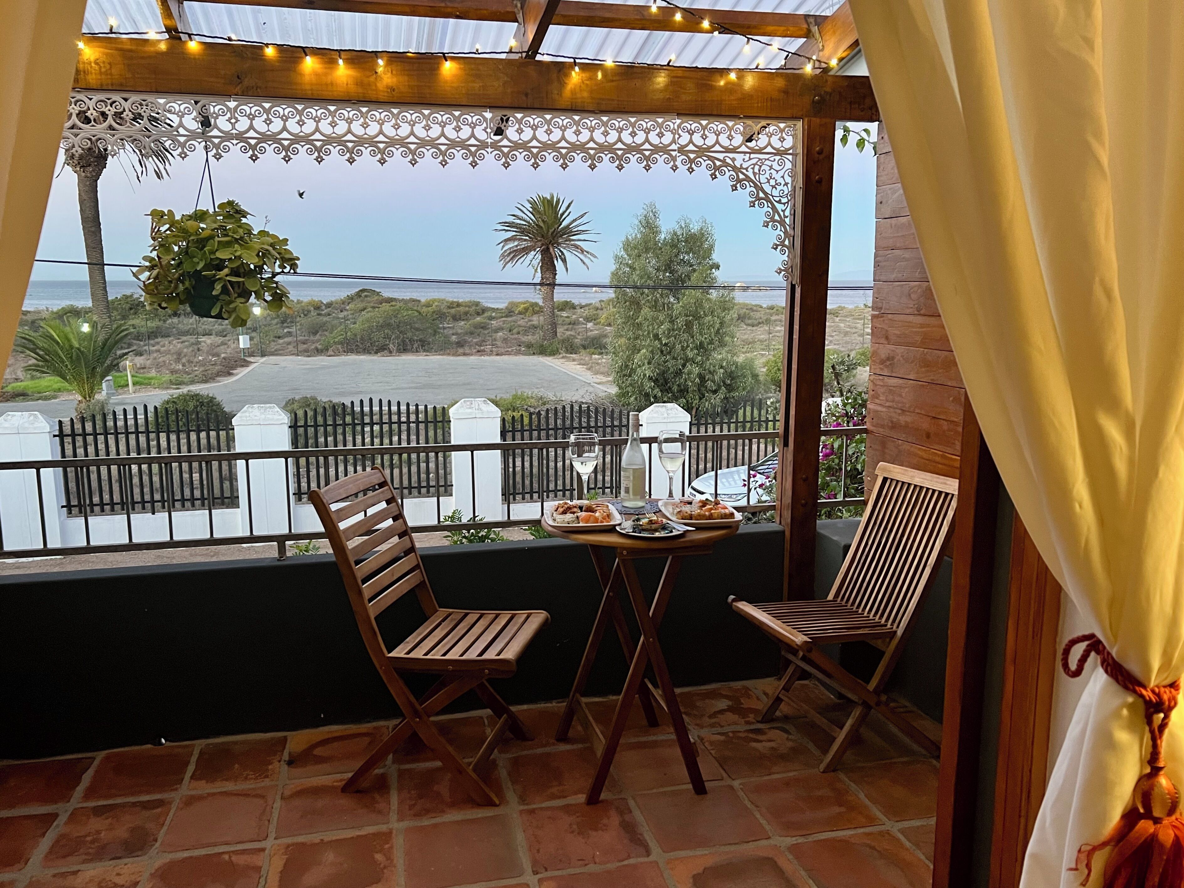 A balcony with two wooden chairs, a table with plates of food and drinks, and a view of a coastal road.