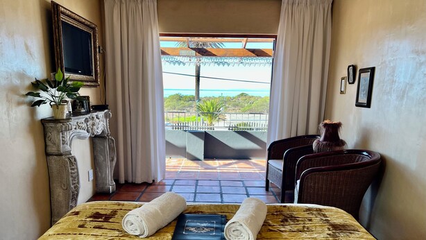 A hotel room with a bed, a desk, a chair, a potted plant, and a view of the outdoors. - Goblin's Inn (St. Helena Bay)