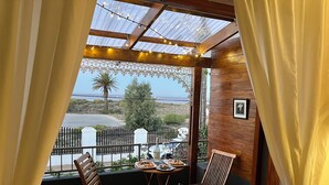 A balcony with two wooden chairs, a small table with a bottle and snacks, and a view of the sea and greenery. - Goblin's Inn (St. Helena Bay)