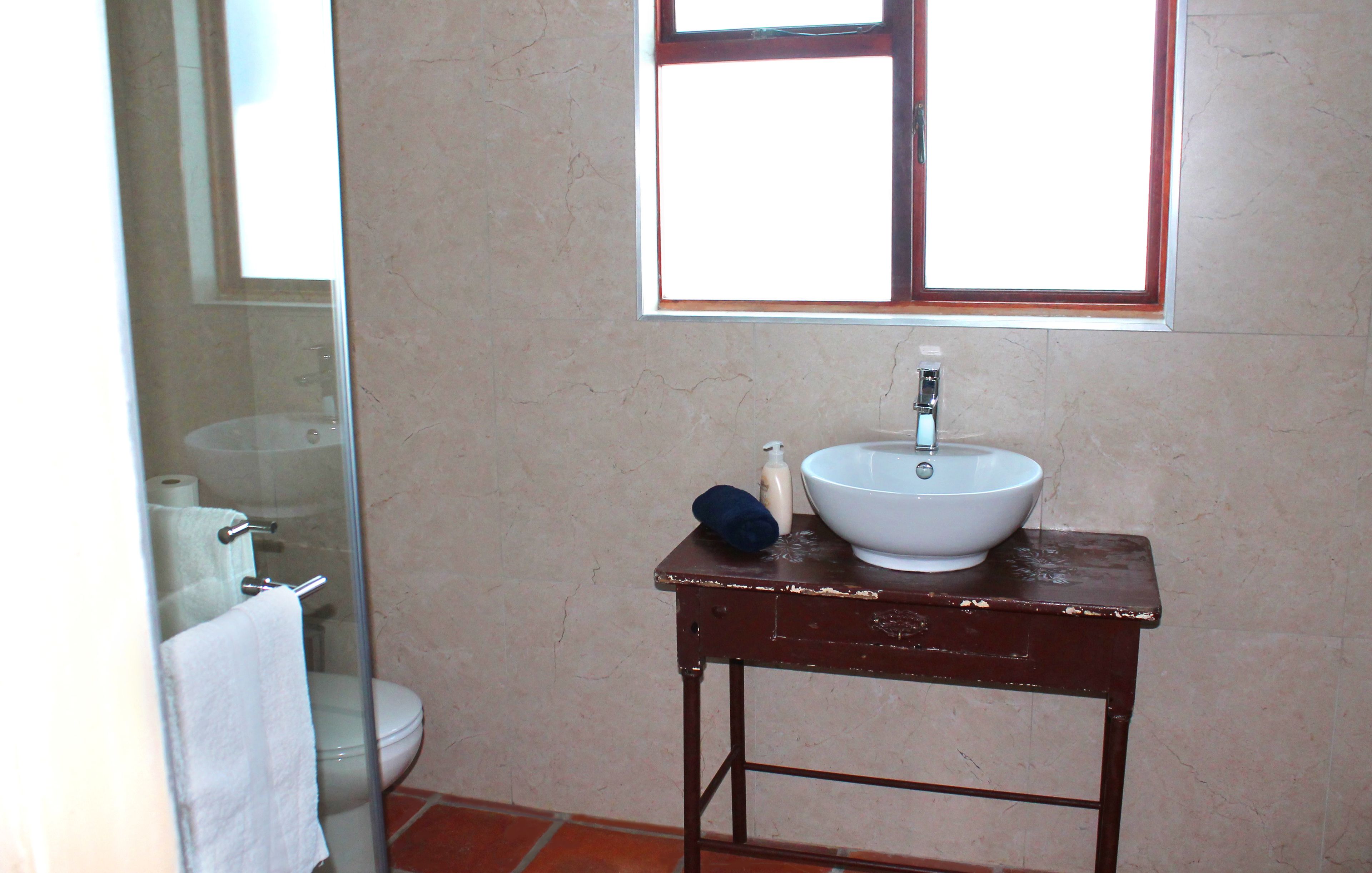 A bathroom with a white sink on a wooden table, a window above the sink, and a glass shower enclosure.