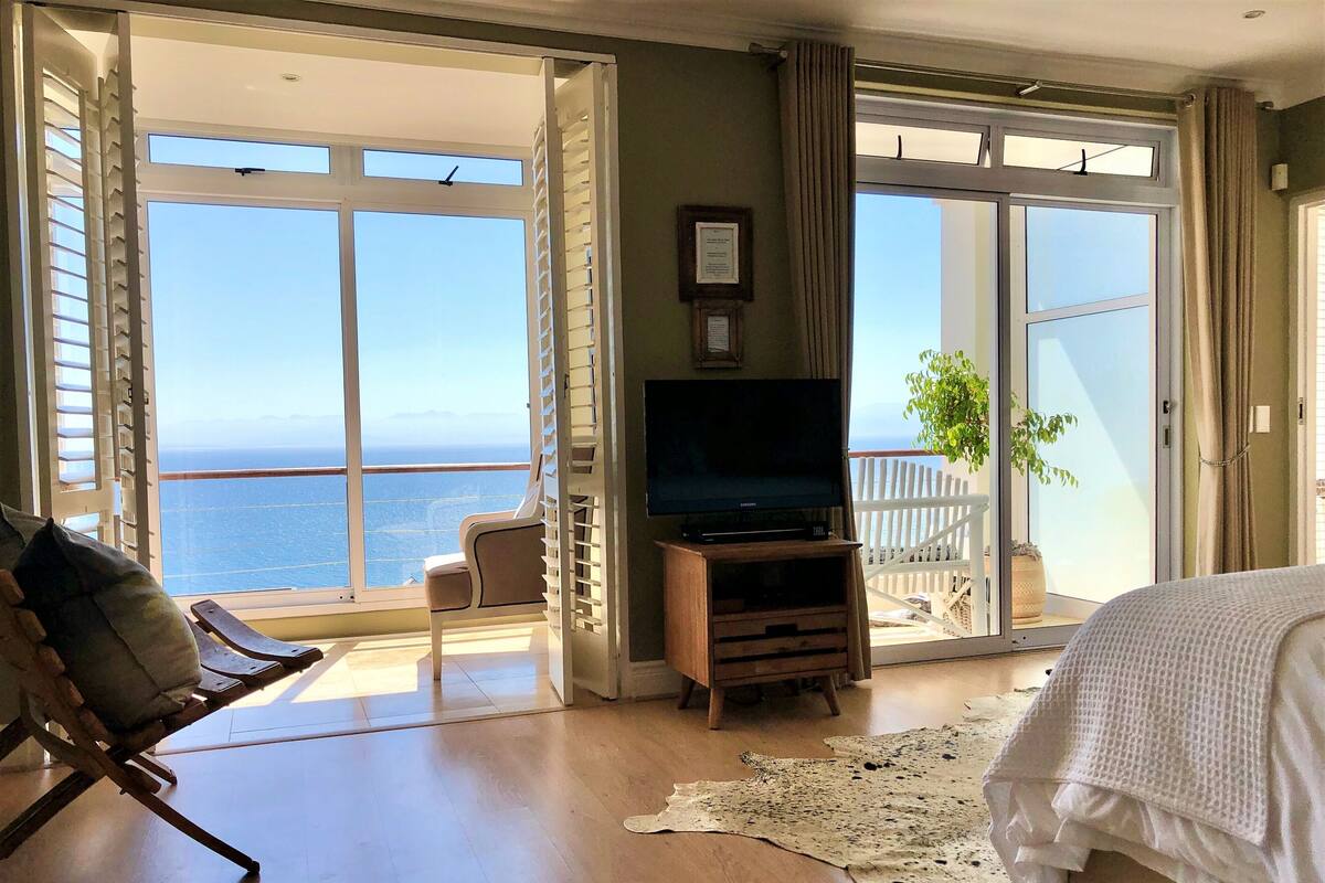 Une pièce dotée d’une grande porte-fenêtre coulissante donnant sur un balcon avec vue sur la mer. On y trouve un meuble TV en bois avec un téléviseur à écran plat, une chaise et un lit recouvert d’une courtepointe blanche.