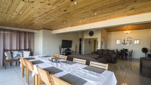 A spacious living area with a wooden ceiling, a dining table set with chairs, a sofa, and a TV mounted on the wall. - Madiolyn (Jeffreys Bay)