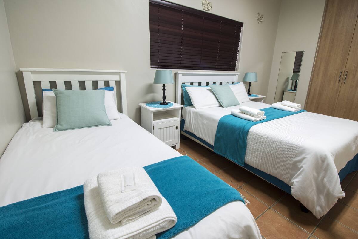 A hotel room with two beds, each with a white headboard, blue bedding, and a nightstand with a lamp.
