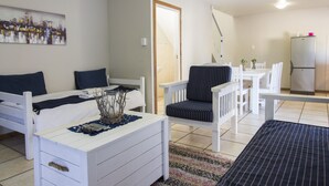 A room with a bed, a desk, a chair, a dining table with chairs, and a refrigerator. - Madiolyn (Jeffreys Bay)