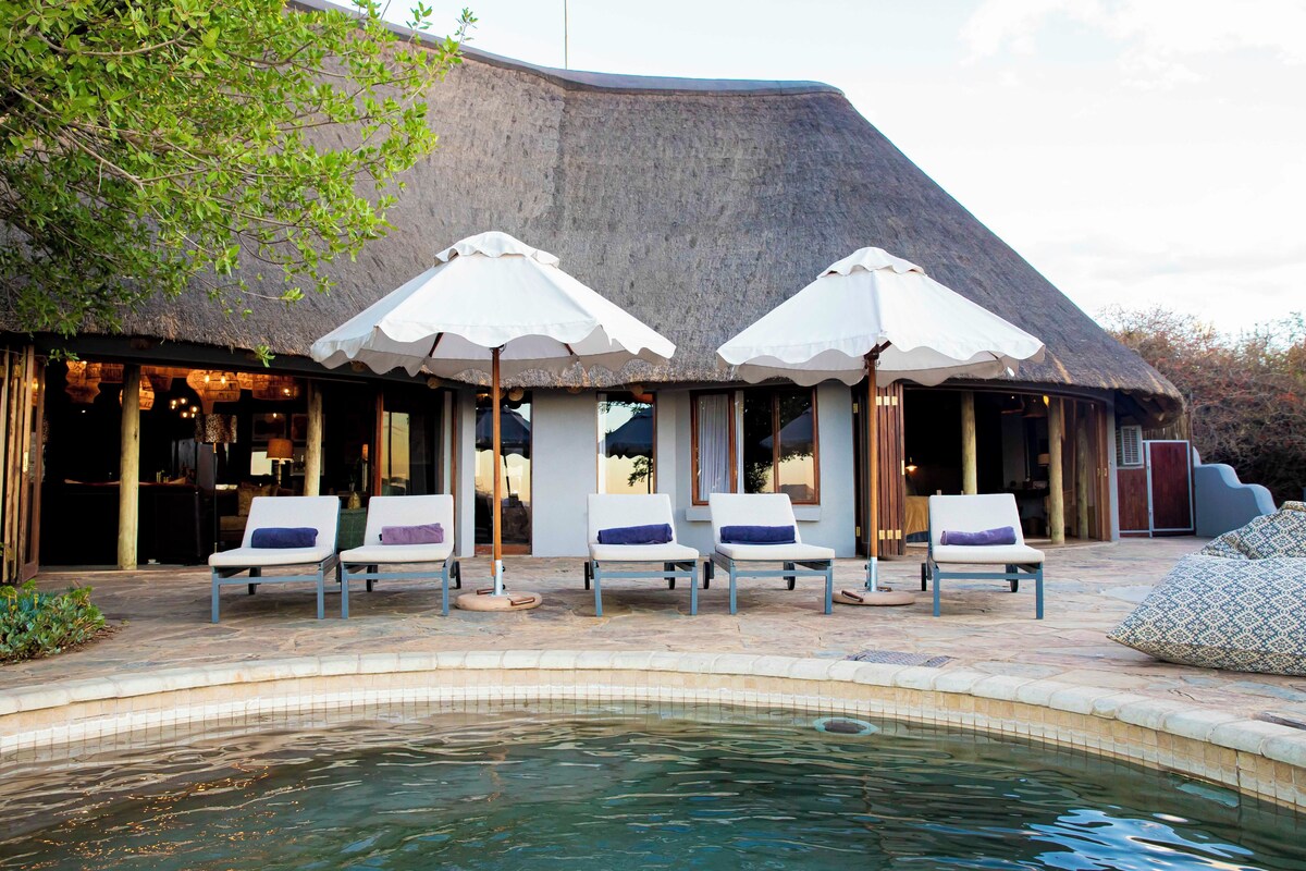 A poolside area with lounge chairs and umbrellas, a pool, and a building with a thatched roof.