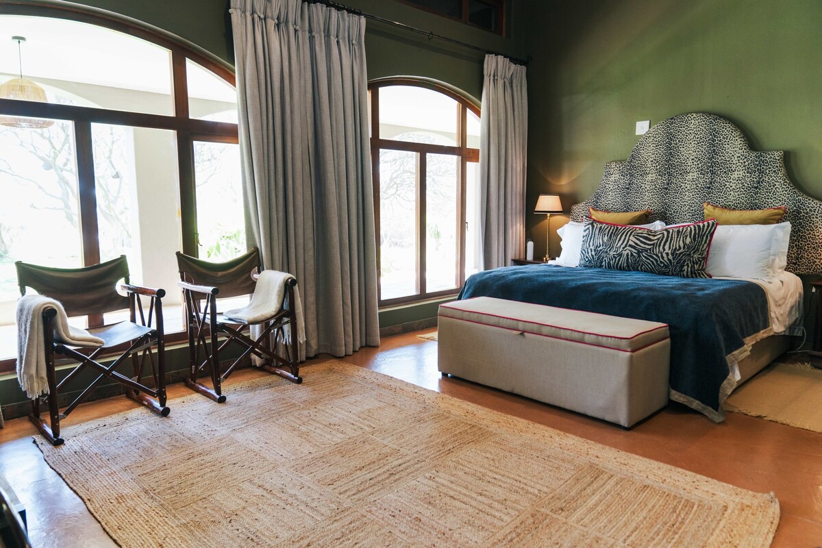 A bedroom with a large bed, a nightstand, a lamp, a chair, a bench, and a window with curtains.