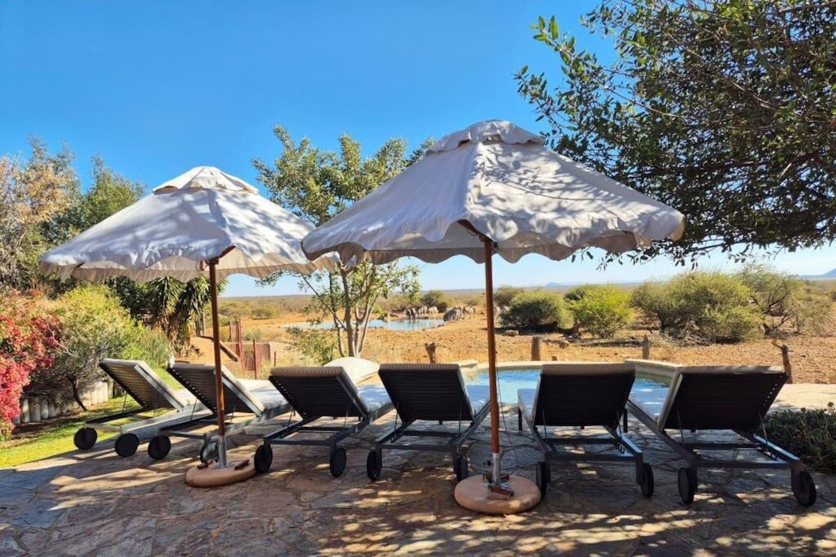A poolside area with lounge chairs and umbrellas, a clear sky, and a natural landscape in the background.