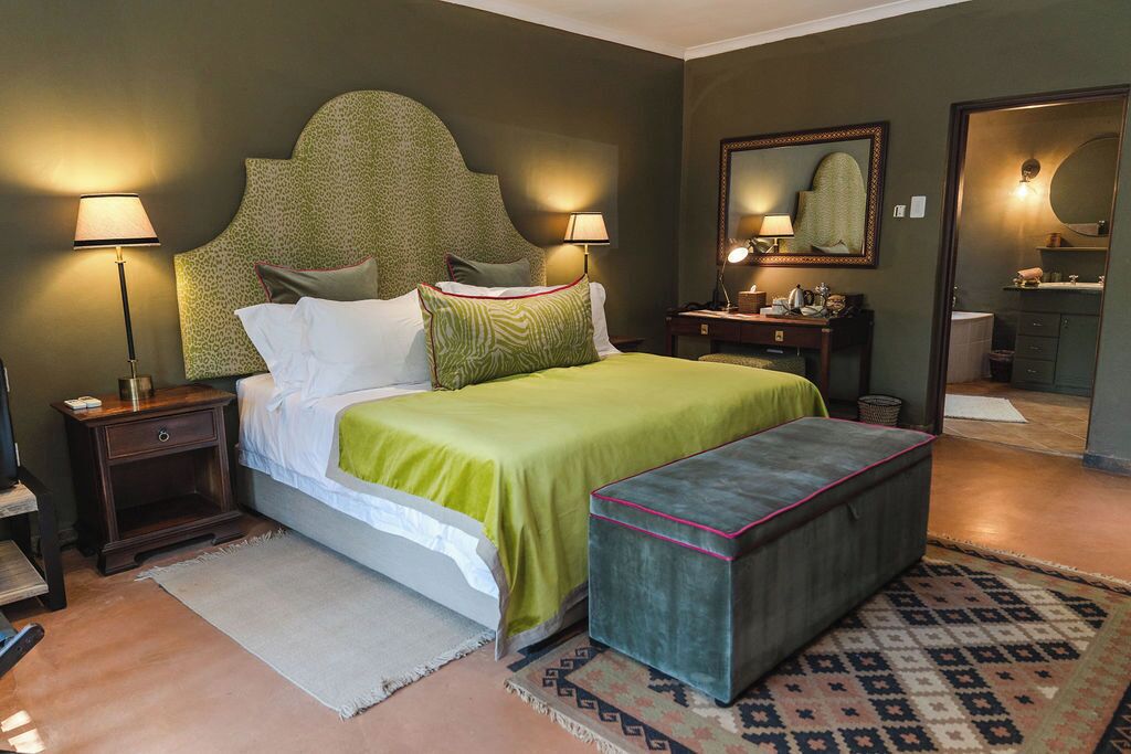 Un dormitorio con cama, mesitas de noche, lámparas, un espejo y un pequeño banco.