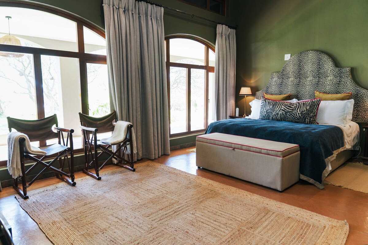 A bedroom with a large bed, a nightstand, a lamp, a chair, a bench, and a window with curtains.