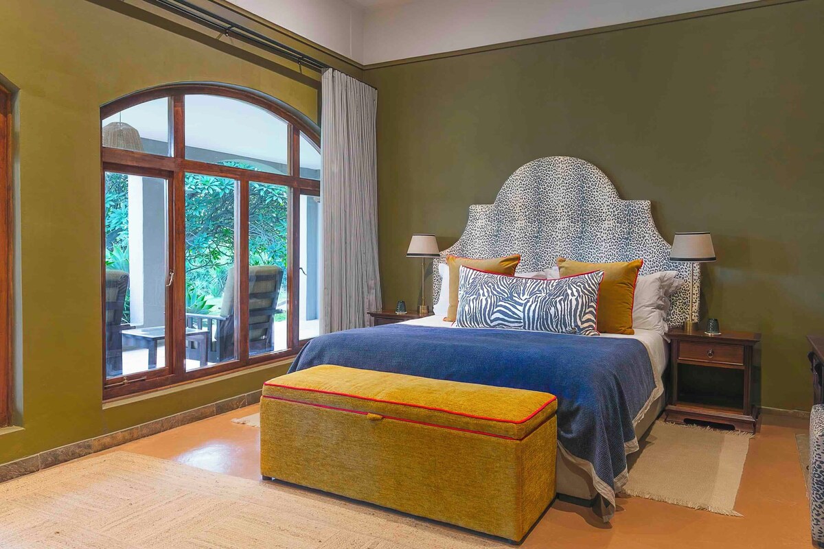 A bedroom with a large window, a bed with patterned pillows, a nightstand, and a small table.