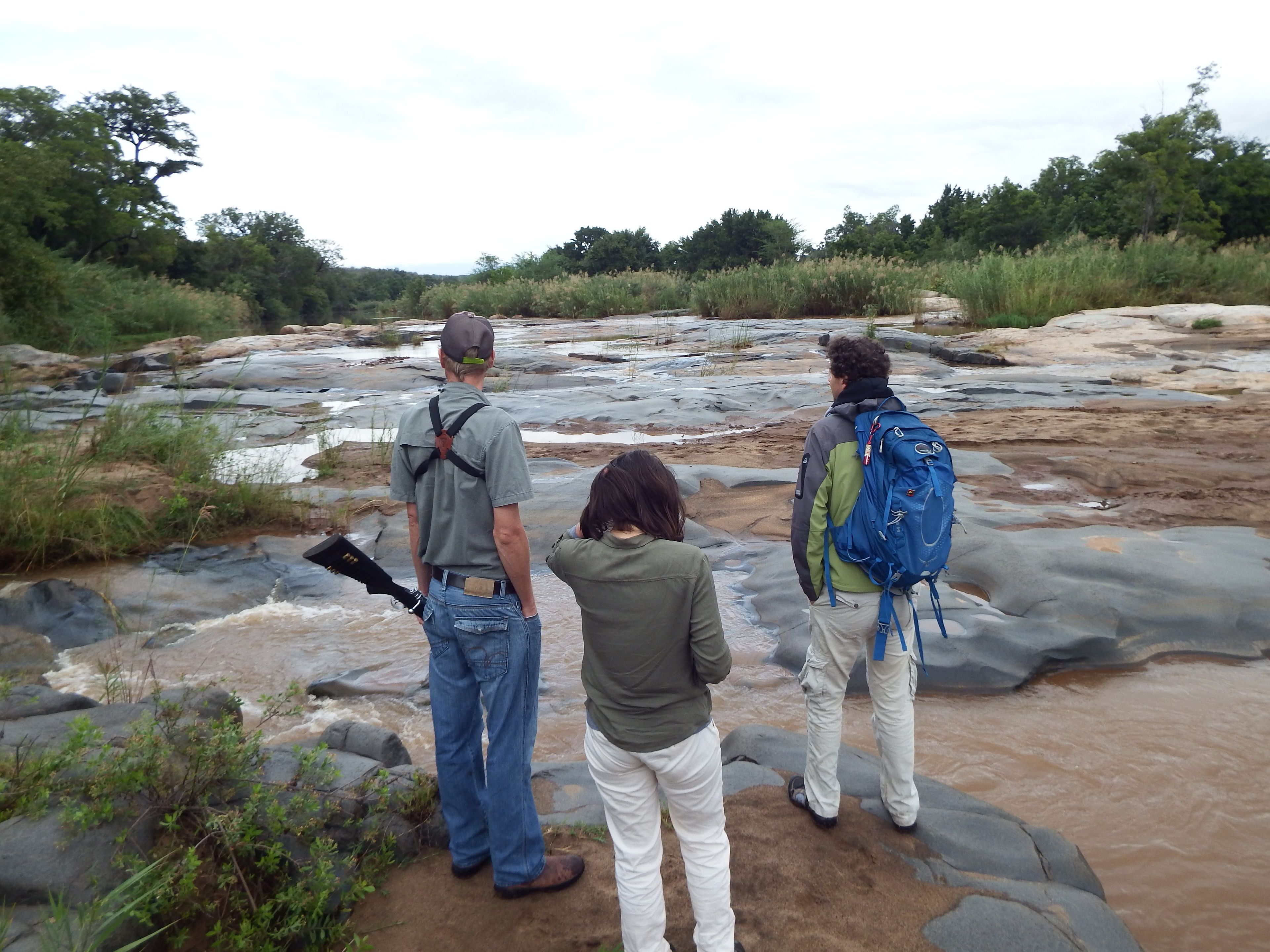 Three people standing on rocky terrain near a flowing river, surrounded by greenery.