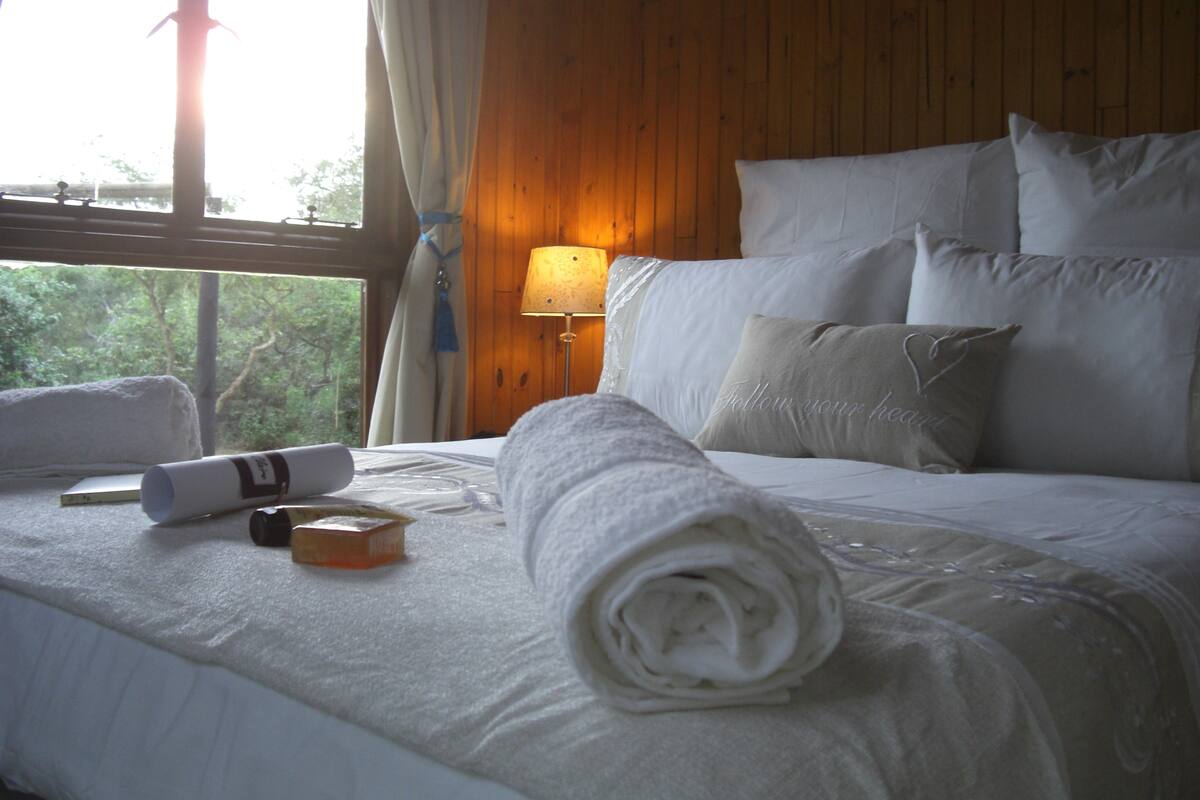 Een net opgemaakt bed met witte lakens en kussens, een opgerolde handdoek en een flesje lotion op het bed.