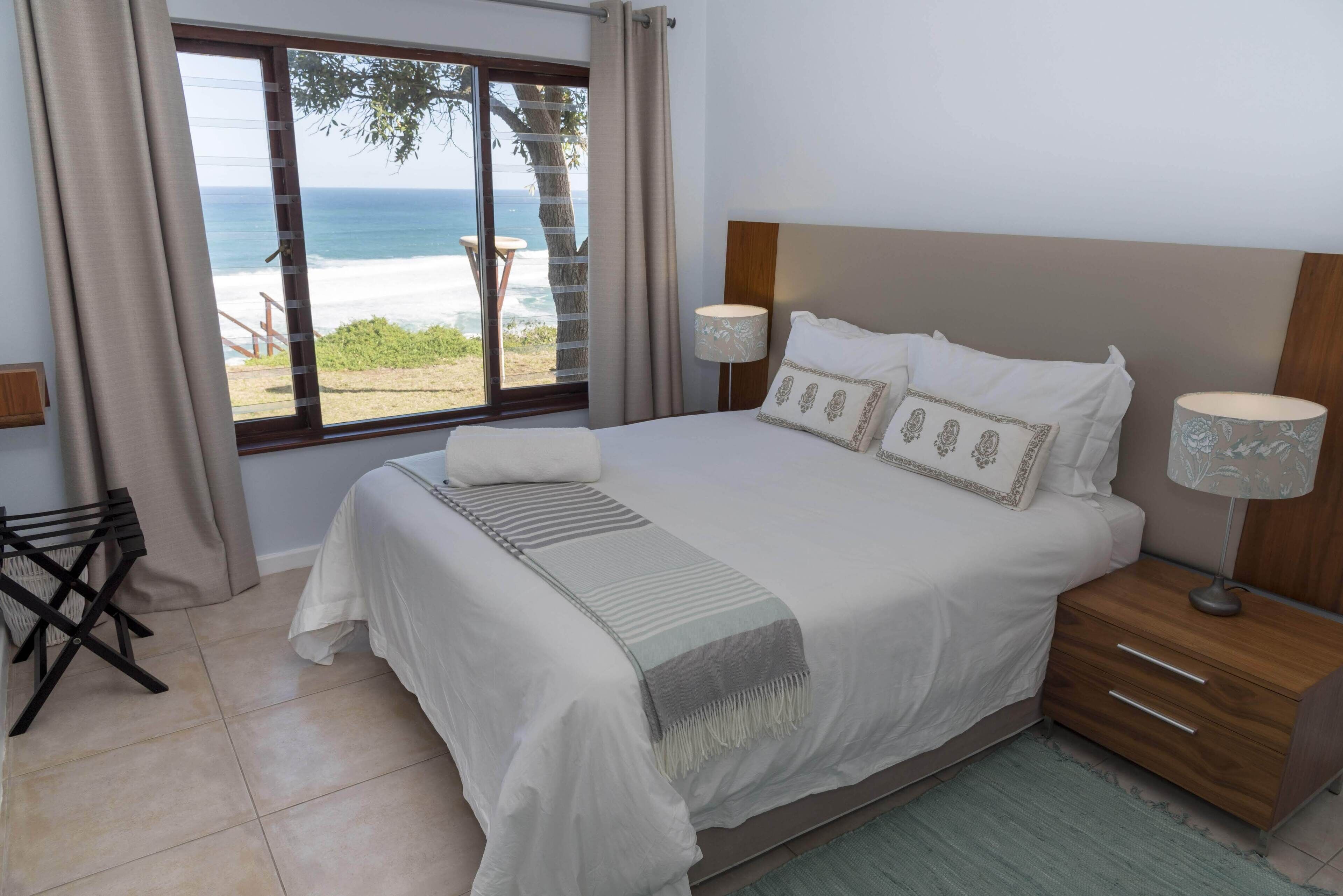 A bedroom with a large bed, a wooden nightstand, a lamp, and a view of the beach and ocean through a window.