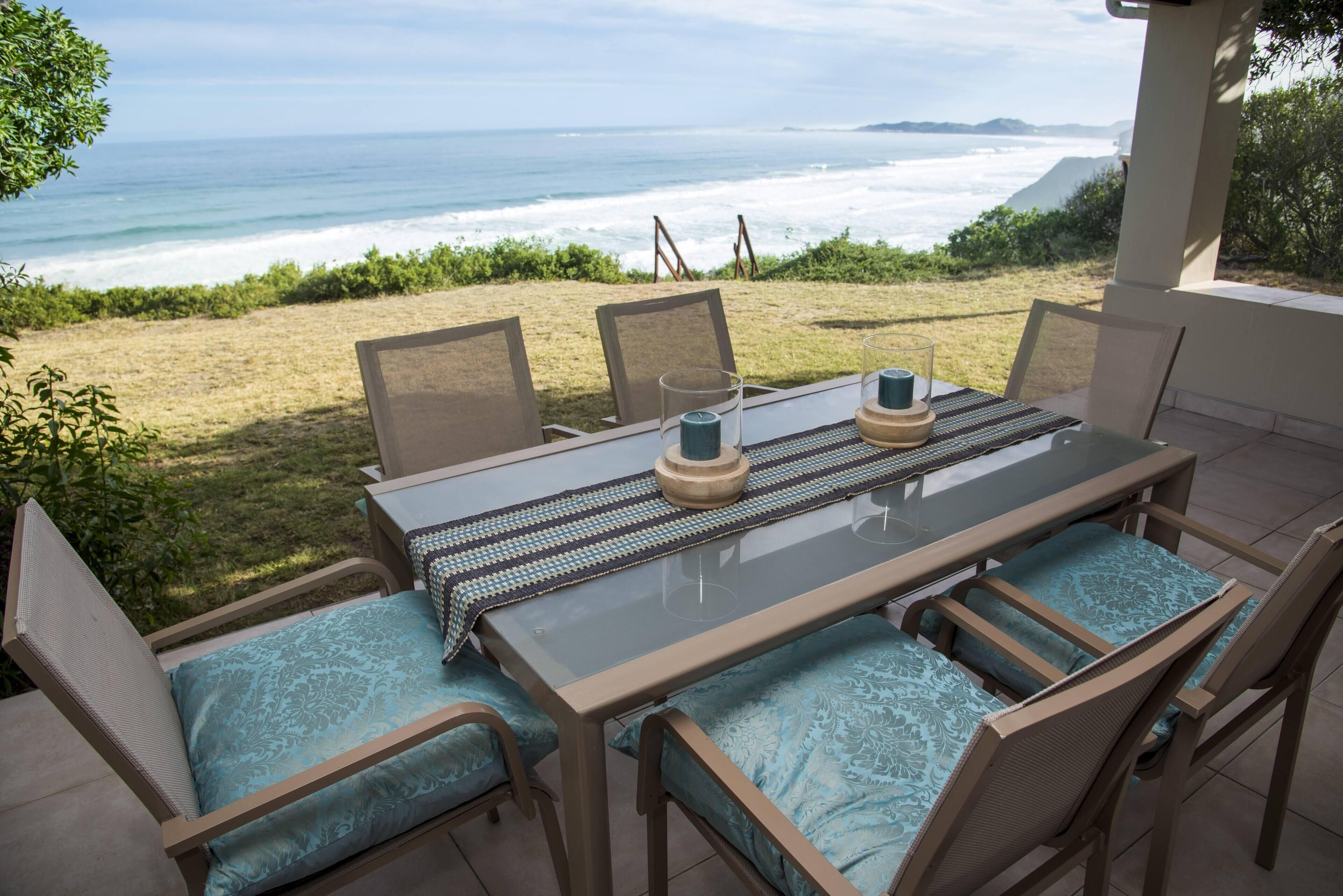 Une terrasse extérieure avec une table et des chaises, donnant sur une plage où l’on peut admirer les vagues et la côte au loin.