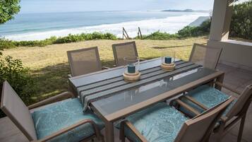 Une terrasse extérieure avec une table et des chaises, donnant sur une plage où l’on peut admirer les vagues et la côte au loin.