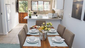 A modern kitchen with white cabinets, a dining area with a wooden table set for four, and a view of the outdoors through large windows.