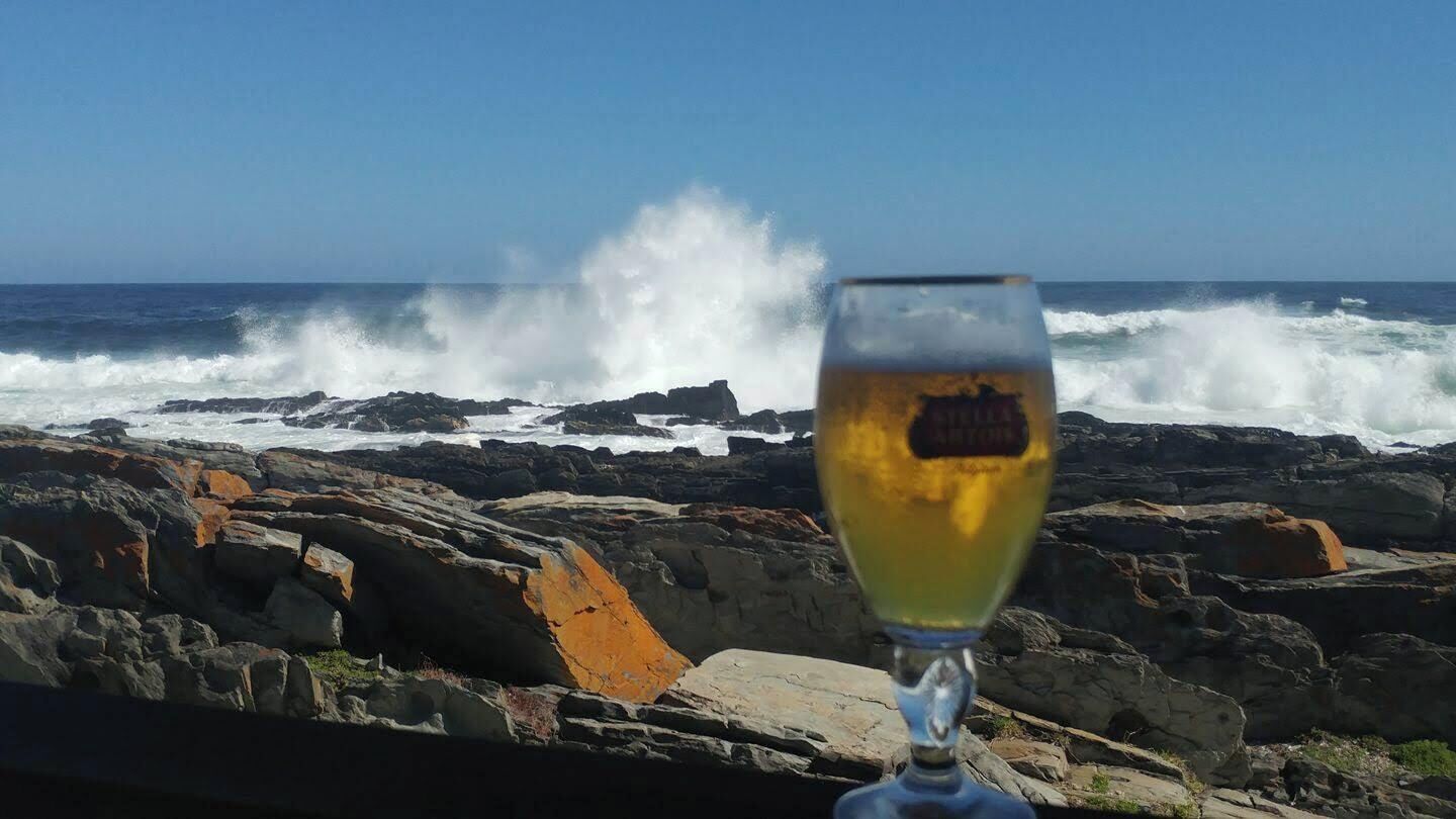 A glass of white wine with a label, placed in front of a rocky coastline with waves crashing against the rocks.