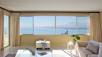 A spacious living room with a large window offering a view of a mountain and the sea. There is a white sofa, a glass coffee table with a magazine, and a small side table with a plant.