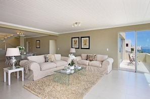 A spacious living room with a beige sofa set, a glass coffee table, and a large window offering a view of the sea. - Bayview Gordon's Bay (Cape Town)
