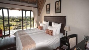 Un grand lit avec du linge de lit blanc et des oreillers à motifs, une tête de lit en bois et une vue sur un paysage luxuriant, visible à travers de grandes fenêtres.