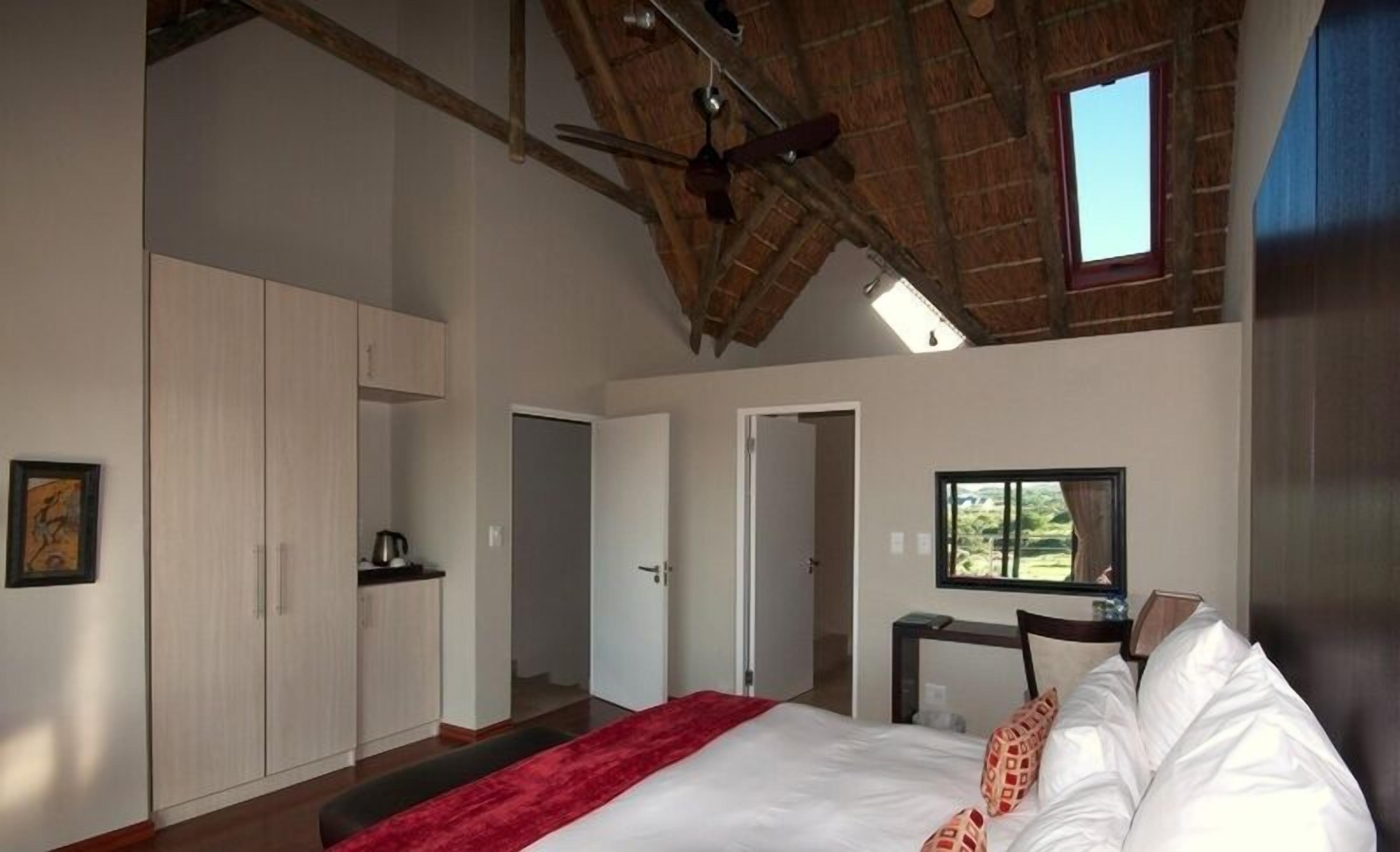 A bedroom with a large bed, a red throw, a ceiling fan, and a view of greenery outside.