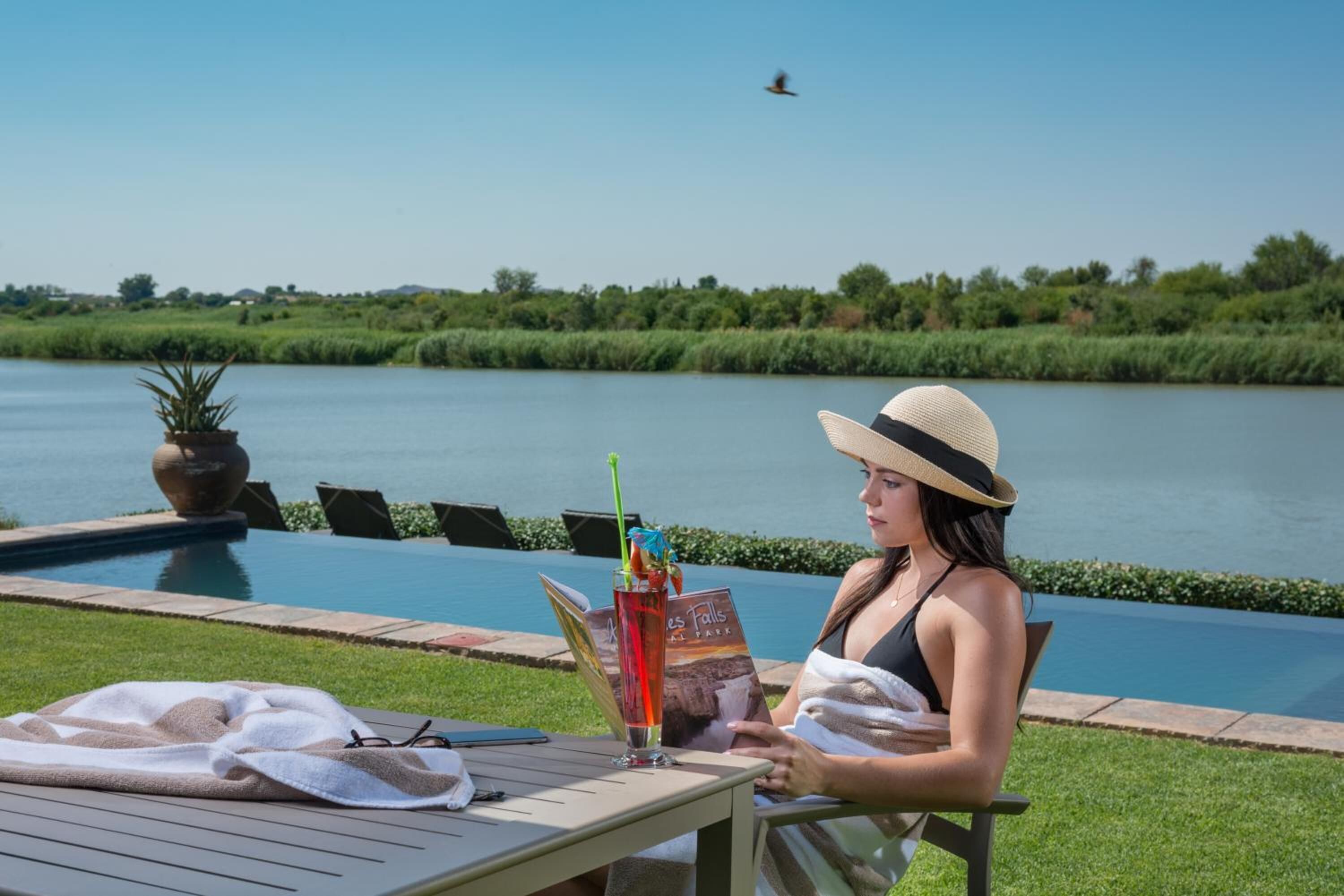 Eine Frau sitzt am Pool mit einem Buch und einem Getränk und blickt auf einen See.