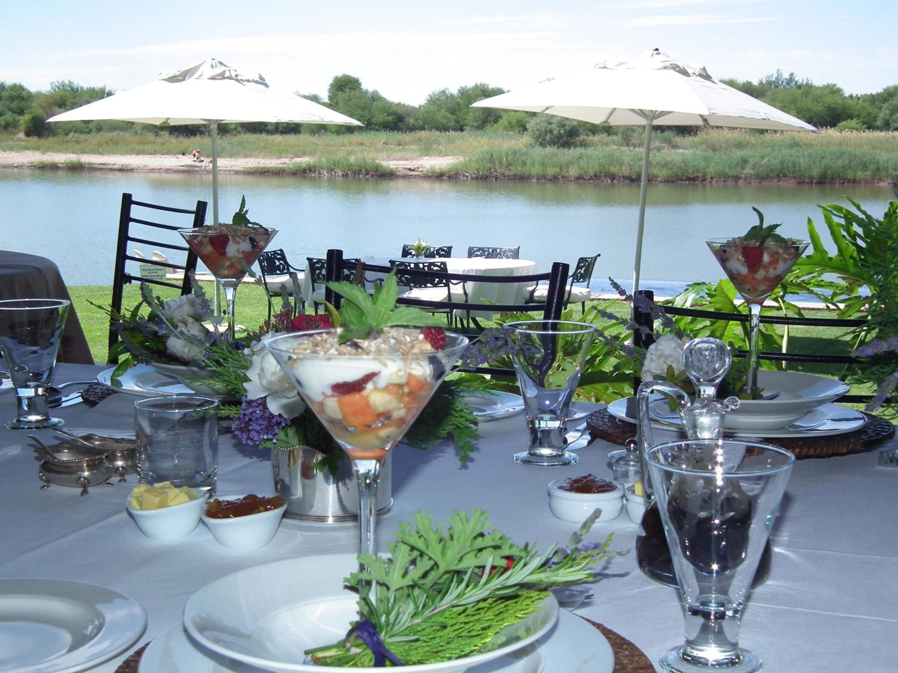 A table set for a meal with a view of a lake, outdoor chairs, and umbrellas.