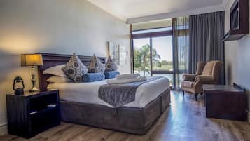 Uma cama grande com almofadas estampadas, uma mesinha de cabeceira com abajur, uma cadeira e vista para o exterior através de grandes janelas.