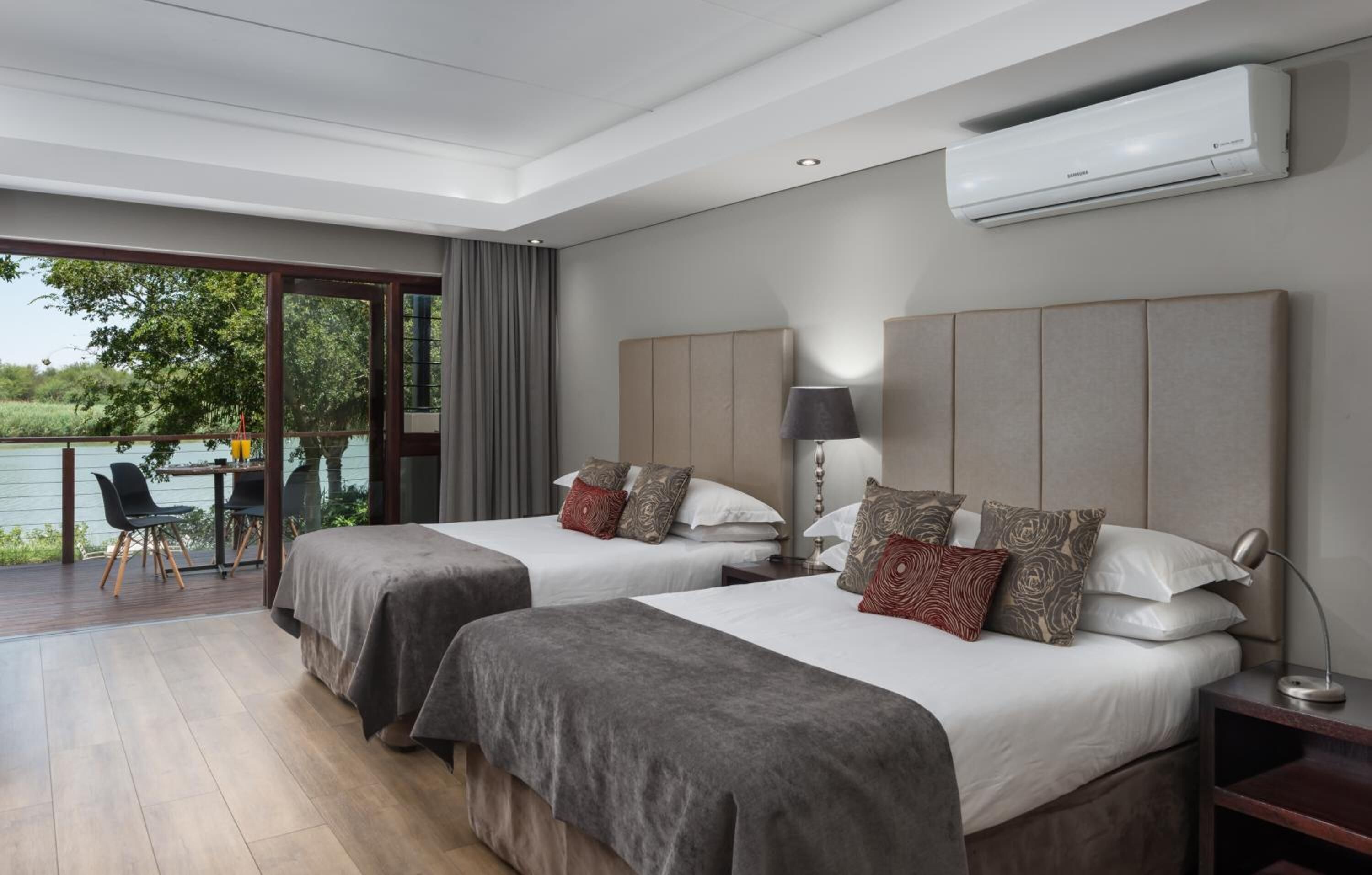 A modern hotel room with two beds, a balcony with a table and chairs, and a view of a lake.