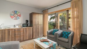 A compact living space with a kitchenette, a sofa set, a coffee table, and a large window with curtains. - Overmeer Guest House (Knysna)