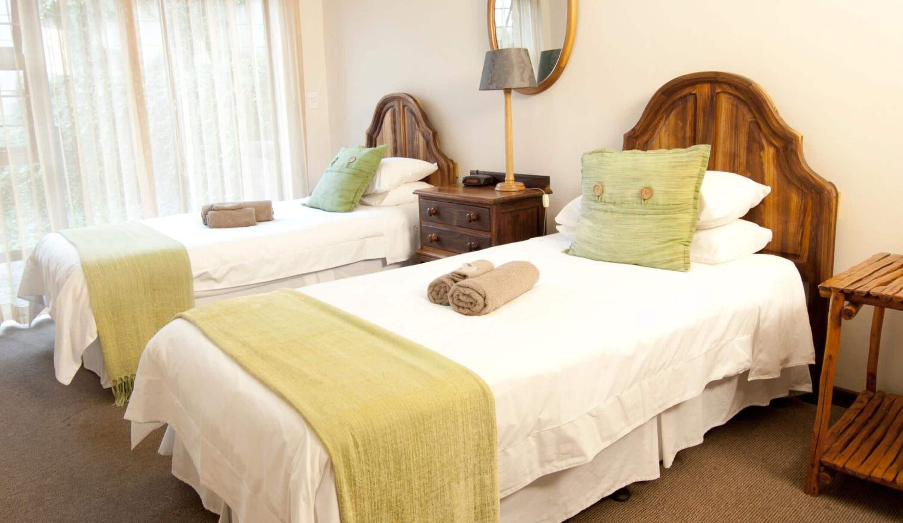 Habitación de hotel con dos camas, cada una con cabecero de madera, ropa de cama blanca y una manta verde. Hay una mesita de noche de madera con una lámpara y un espejo encima.