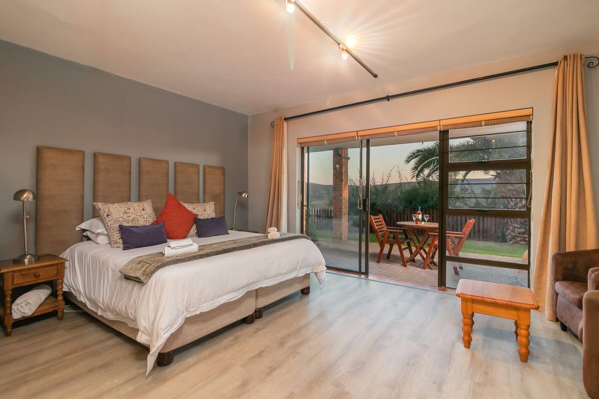 A bedroom with a large bed, a wooden nightstand, a bedside table, a wooden bench, and a sliding glass door leading to an outdoor patio with a table and chairs.