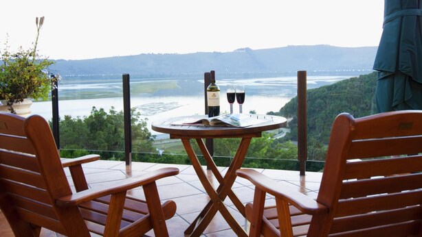 A wooden table with a bottle of wine and two glasses on a balcony overlooking a river and hills. - Overmeer Guest House (Knysna)