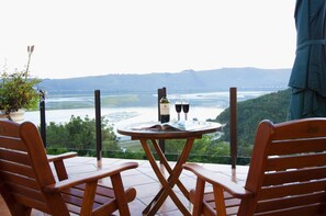 A wooden table with a bottle of wine and two glasses on a balcony overlooking a river and hills. - Overmeer Guest House (Knysna)