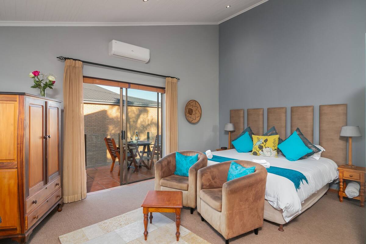 A bedroom with a bed, two armchairs, a wooden cabinet, and a sliding glass door leading to a patio.