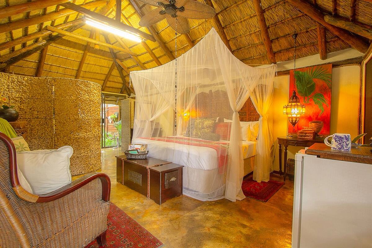 A room with a bed under a mosquito net, a wicker chair, a small table, and a refrigerator.