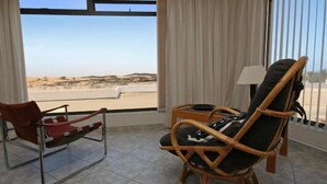 Une pièce avec vue sur un paysage désertique, grâce à de grandes fenêtres, comprenant un fauteuil rouge, un fauteuil en osier et une petite table avec une lampe.