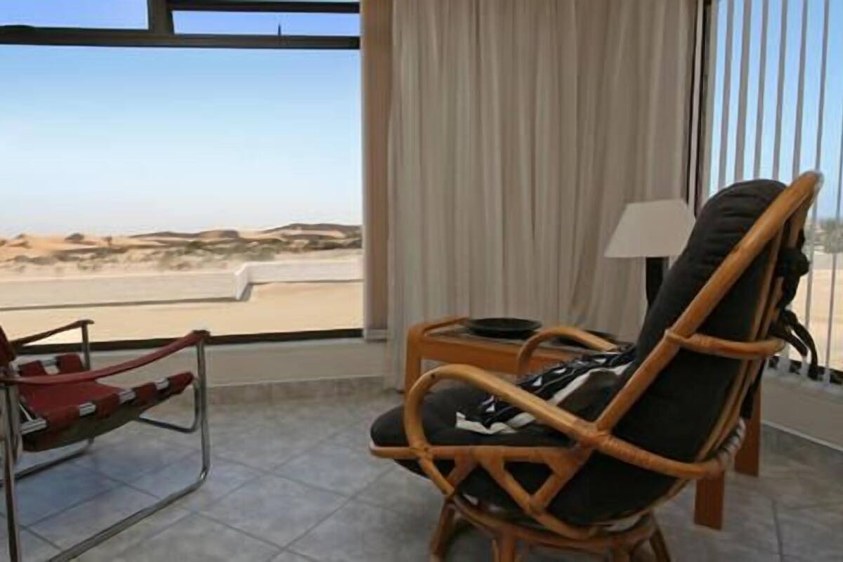 A room with a view of a desert landscape through large windows, a red chair, a wicker chair, and a small table with a lamp.