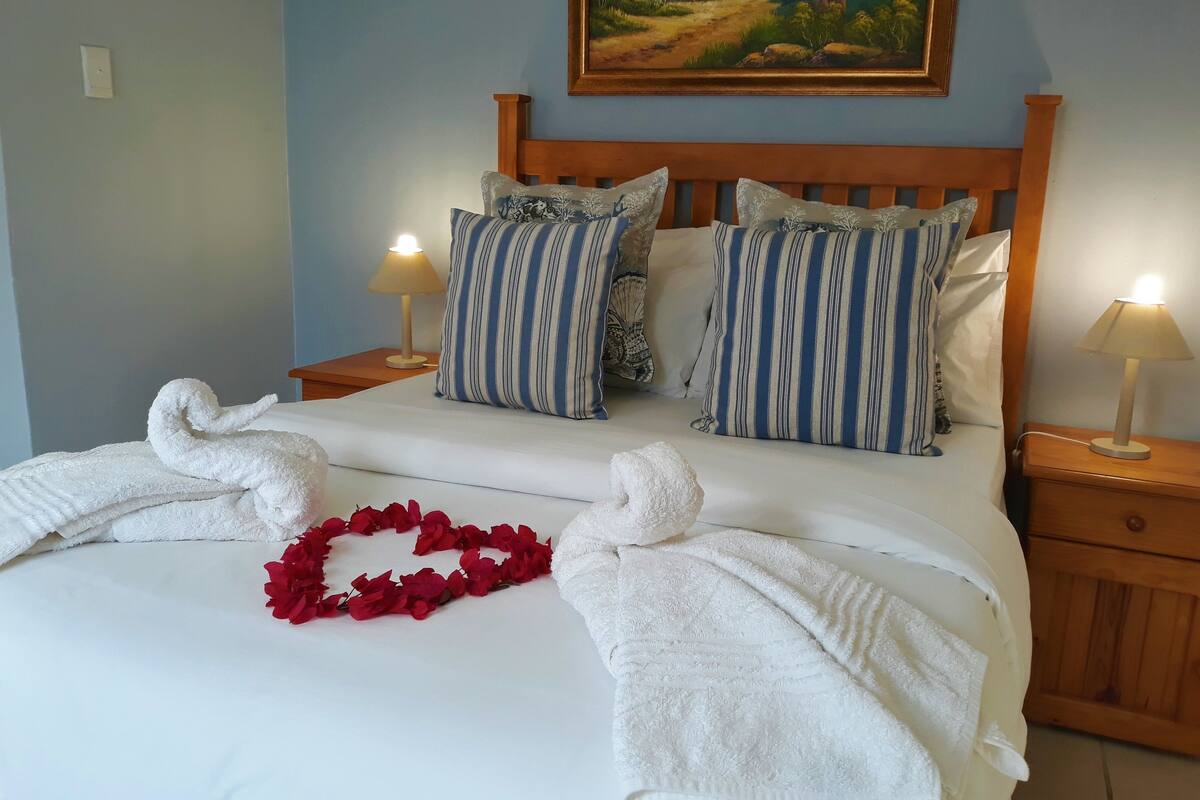 Una cama bien hecha con sábanas blancas, almohadas a rayas y un arco de flores rojas. Hay dos lámparas de noche y un cuadro sobre la cama.
