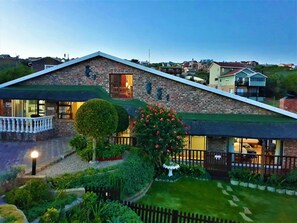 A two-story house with a brick exterior, a green roof, and a well-maintained garden with a round hedge and flowering plants.