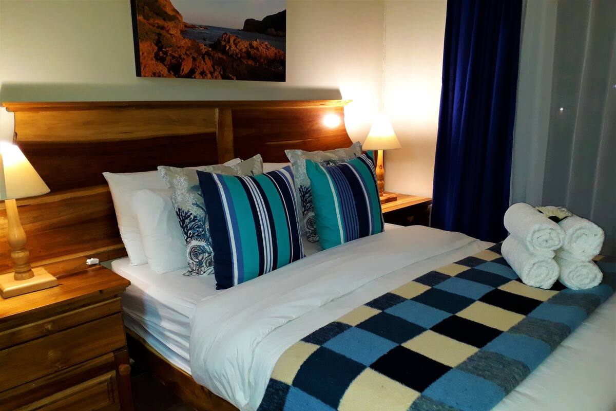 Habitación de hotel con una cama, dos lámparas de noche, un cabecero de madera, una colcha a cuadros y un cuadro de una escena costera.