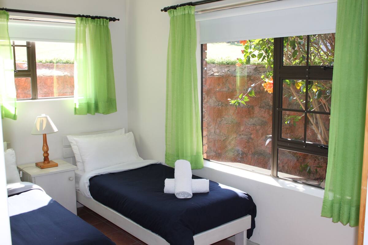 Un dormitorio con una cama, una mesita de noche con lámpara y una ventana con cortinas verdes.