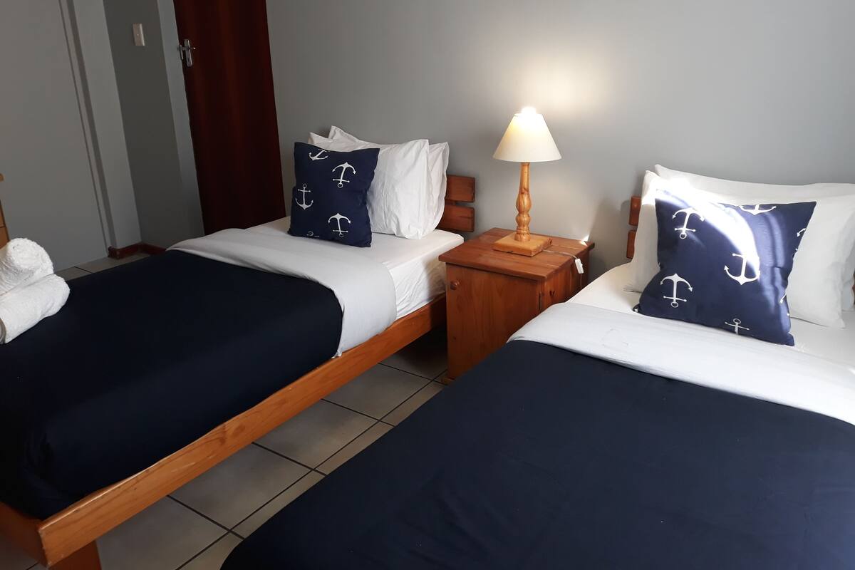 Habitación de hotel con dos camas, cada una con ropa de cama azul marino y almohadas blancas con estampados de anclas. Entre las camas, hay una mesita de noche de madera con una lámpara.