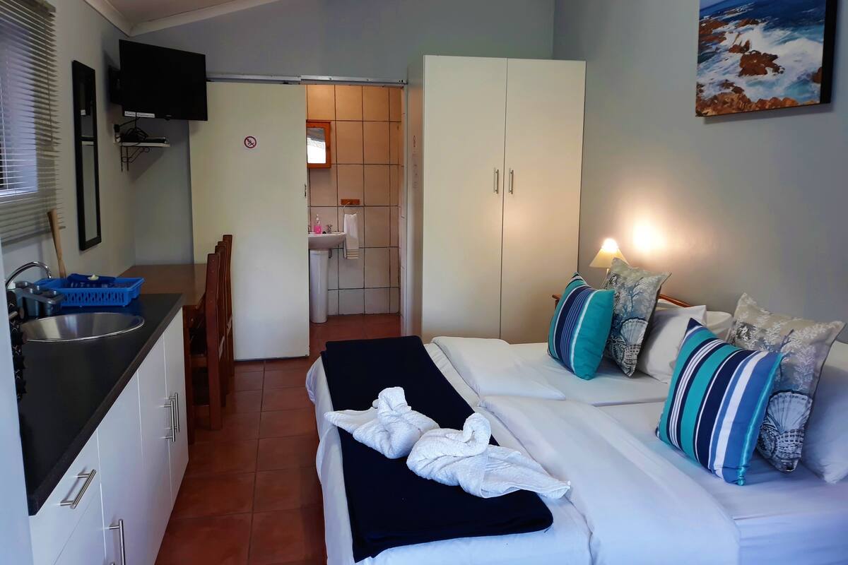 Una habitación de hotel compacta con kitchenette, una cama con colcha blanca y negra, y un cuadro con un paisaje marino.